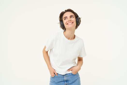 Image of modern girl in wireless headphones, listens to music in earphones, smiles and looks happy, poses in casual clothes over white background.