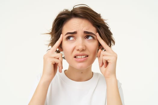 Image of woman touches her head temples, looks concerned, discomfort, has headache, tries to remember, thinking hard, standing over white studio background.