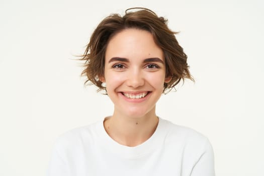 Close up shot of brunette girl with short hairstyle with genuine emotions, smiling and looking happy, standing over white background. Copy space