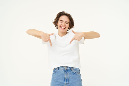 Image of beautiful, happy brunette woman, pointing fingers down, showing advertisement, standing over white studio background. Copy space