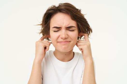 Young woman shuts her ears from loud noise, hears siren, distressed by shouting neighbours, standing over white background.