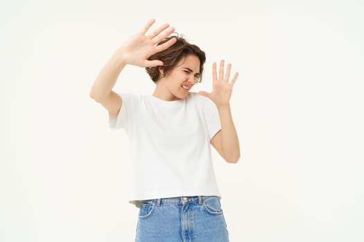 Portrait of woman raising hands in defense and blocking face from something, standing over white background.