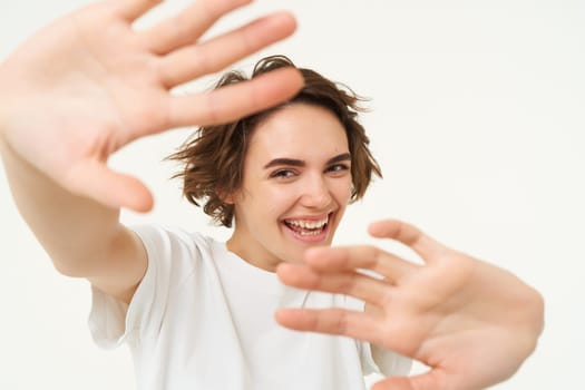 Portrait of happy, laughing young woman, blocks herself from camera, looking joyful, stretching her hands forward, posing over white background. Copy space