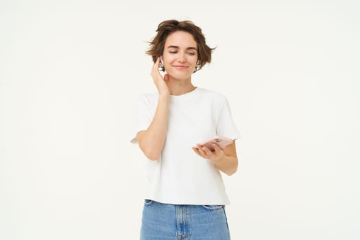 Portrait of woman puts on wireless headphones and holds smartphone, listens music, stands over white background.