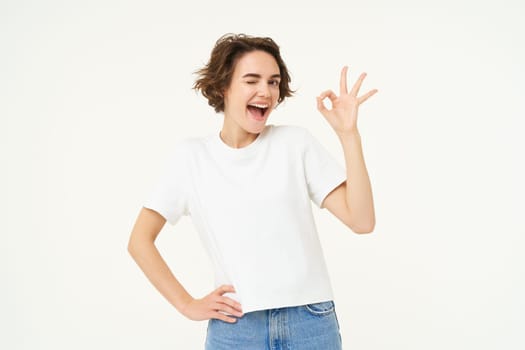 Portrait of cute excited woman, winks at you, shows okay, ok gesture, poses over white studio background.