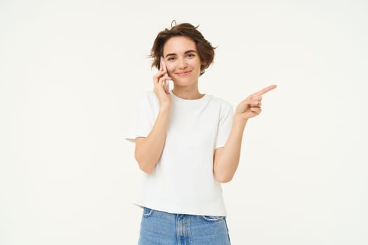 Portrait of young woman talking on mobile phone, using smartphone and pointing finger right, showing direction, banner advertisement, studio background.
