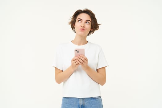 Portrait of woman thinking, using smartphone and looking thoughtful, making choice, deciding, standing isolated over white background.