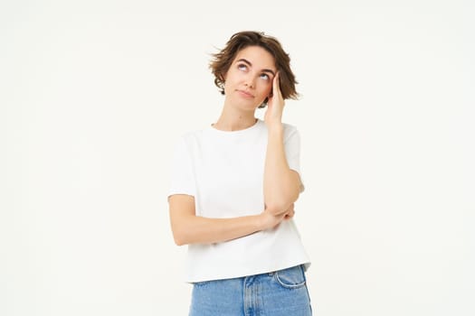 Portrait of puzzled woman, looking confused, staring up at ceiling, touches her head, standing tired against white background.