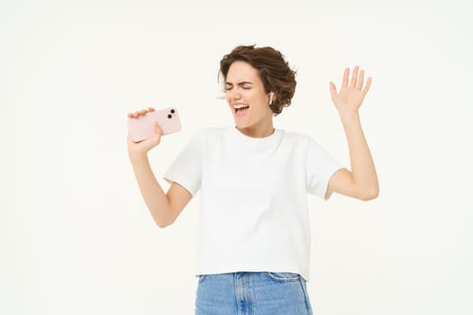 Portrait of carefree girl singing in wireless headphones into smartphone microphone, playing music game on mobile app, standing over white background.