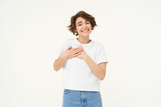 Portrait of woman expressing care, self-love and caring emotions, holds hands on chest, looking with smile at camera, isolated over white background.