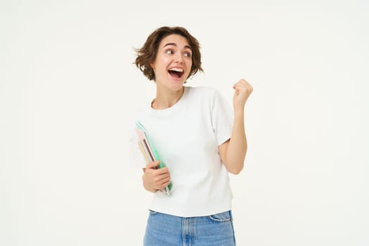 Enthusiastic brunette woman makes fist pump, holds documents and notes, looks thrilled and happy, winning, triumphing, posing over white background.