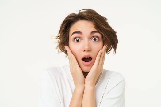 Portrait of girl with surprised face, makes shocked expression, stands over white background. Emotions and people concept