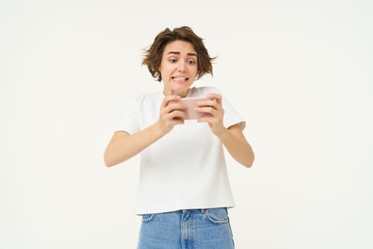 Portrait of excited woman playing smartphone games, looking intense at mobile phone screen, standing against white studio background. Copy space