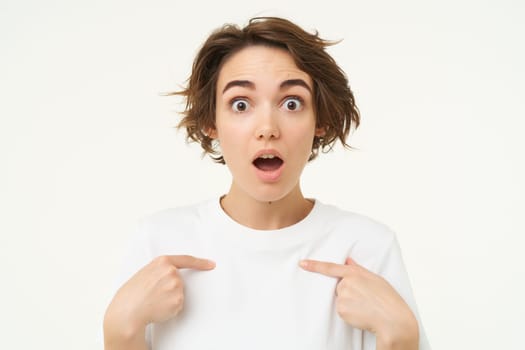 Close up portrait of girl with surprised face, points at herself, looks shocked, stands over white background.