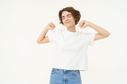 Portrait of smiling, pleased woman stretching hands, waking up after sleep, standing over white background. Copy space