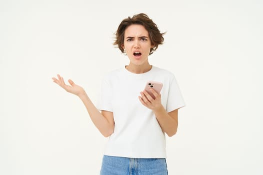Portrait of frustrated woman with mobile phone, looking shocked and upset, complaining, using smartphone, standing over white background.