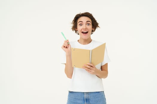 Enthusiastic, creative young woman raises pen, eureka pose, found solution, writing great idea in notebook or planner, stands over white background.