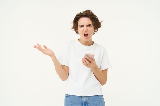 Portrait of frustrated woman with mobile phone, looking shocked and upset, complaining, using smartphone, standing over white background.