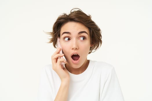 Portrait of amazed brunette girl looks surprised while talks on mobile phone, answers a call, amazed by big news, gasping shocked, standing over white background.