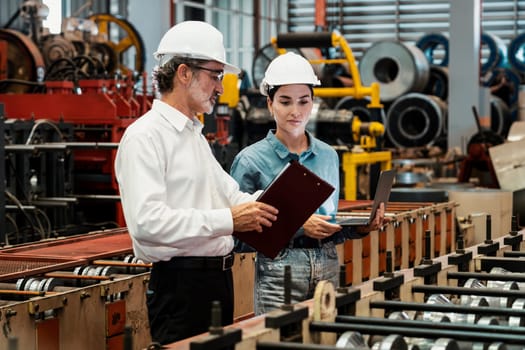 Factory engineer manager with assistant using laptop to conduct inspection of steel industrial machine, exemplifying leadership as machinery engineering inspection supervisor in metalwork manufacture.