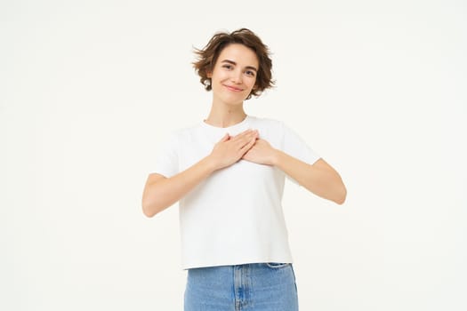 Portrait of brunette woman with hands on chest, looks with care and love, express gratitude, self-care and heart-warming feelings, stands over white background.
