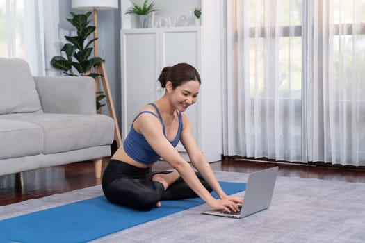 Asian woman in sportswear doing yoga exercise on fitness mat as her home workout training routine. Healthy body care lifestyle woman watching online yoga video on laptop. Vigorous