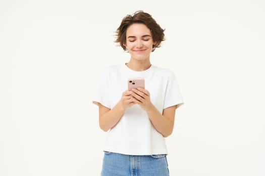 Portrait of young brunette woman texting, sending a message on telephone, using mobile phone and smiling, standing over white studio background.