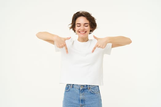 Image of beautiful, happy brunette woman, pointing fingers down, showing advertisement, standing over white studio background. Copy space