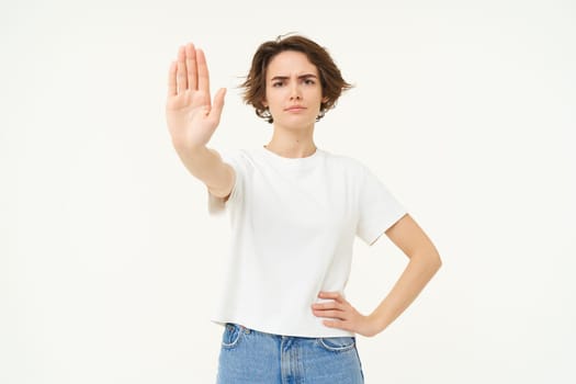 Portrait of serious, confident young woman extends one hand, shows stop, disapproval gesture, looking determined to block way, isolated over white background.
