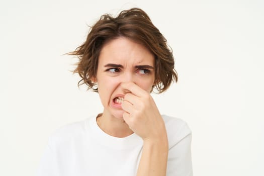 Woman shows dislike, shuts her nose from reeking bad smell, something stinks, stands over white background. Copy space