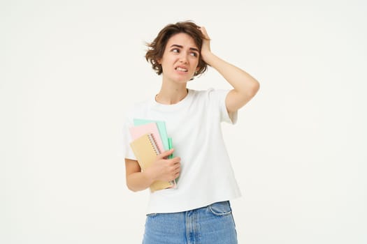 Brunette girl looks troubled, student with problems, holds notebooks and homework with confused face expression, white studio background.