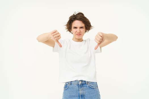 Portrait of disappointed young woman, female client shows dislike gesture, thumbs down, stands over white background.