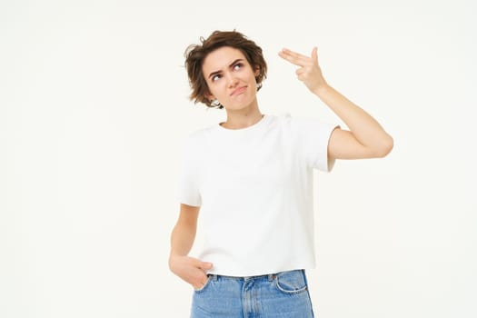 Portrait of stressed and annoyed woman, pointing fingers at forehead, shooting gesture, standing fed up against white background.