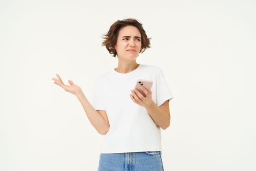 Portrait of woman with smartphone standing confused, puzzled, cant understand something online, using mobile phone, posing over white studio background.