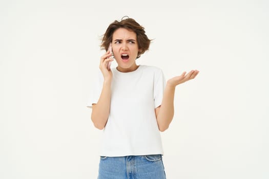Portrait of angry lady talking on phone, frustrated woman shouting at her mobile telephone, standing over white background.