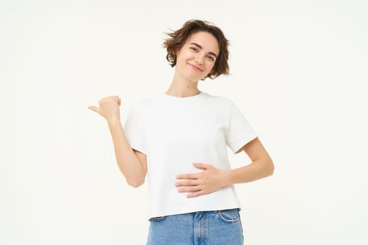 Portrait of smiling, pleased brunette woman, touches belly and points left, shows banner, recommends painkiller, pleased by medication effect on her stomach, stands over white background.