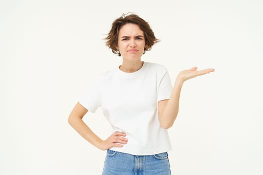 So what. Frustrated, upset young woman, raises one hand and shrugs, looking confused, cant understand something, standing over white background.