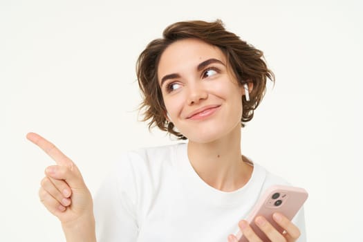 Woman in wireless headphones, holding smartphone, pointing left and smiling, listening to podcast or music, white background.