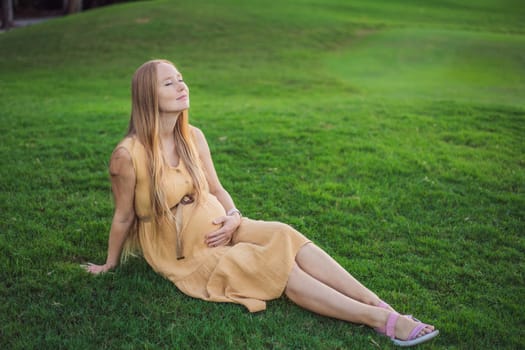 Happy young pregnant model sitting on grass lawn and looking at setting sun in park. Smiling future mom expecting baby. Maternity concept. Copy space.