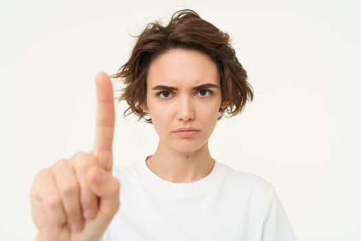 Close up of serious young woman shows one finger, stop, taboo gesture, disapprove something, blocking, saying one rule, standing over white background.