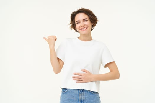 Happy and healthy young woman, pointing left, showing advertisement, touching her belly, feeling better after medication, standing over white background.