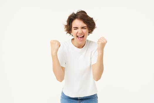 Excited brunette girl, student winning prize, celebrating victory, triumphing, standing in white t-shirt and jeans over white background.