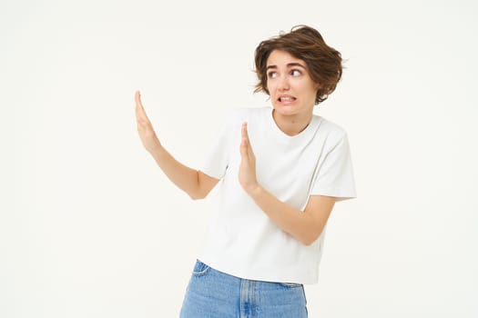 Portrait of awkward, displeased woman shows hand block gesture to the left, avoiding something, refusing, standing over white background.