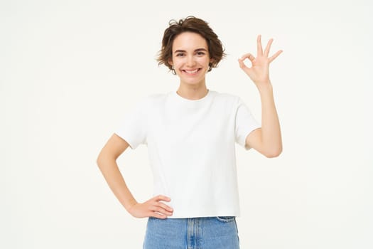 Portrait of woman smiling, showing okay sign with confidence, gives approval, recommends smth good, stands over white background.
