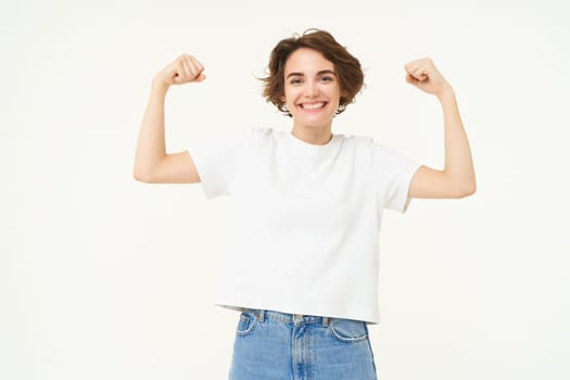 Portrait of happy and confident girl shows muscles, flexing biceps and smiling, posing over white background. People and sports concept