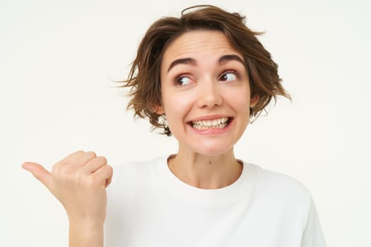 Image of excited brunette woman with short haircut, pointing left, smiling and looking happy, showing you advertisement, standing over white background.