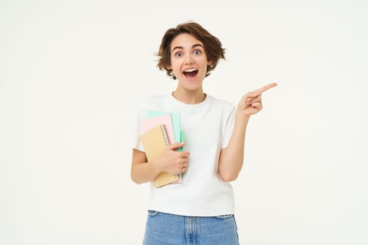 Portrait of brunette woman laughing, student with notebooks pointing at upper right corner, showing banner or advertisement, standing over white studio background.