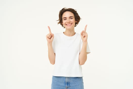 Image of happy, beautiful modern woman, pointing fingers up, showing advertisement on top, smiling, standing over white background.