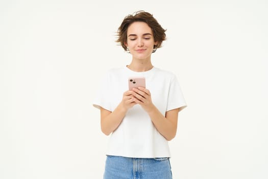 Portrait of brunette woman using smartphone, standing with mobile phone, texting, placing an order, browsing social media in telephone, standing over white background.