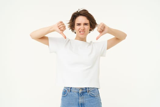 Portrait of disappointed young woman, student shows thumbs down from dislike, disapprove something bad, standing over white background.